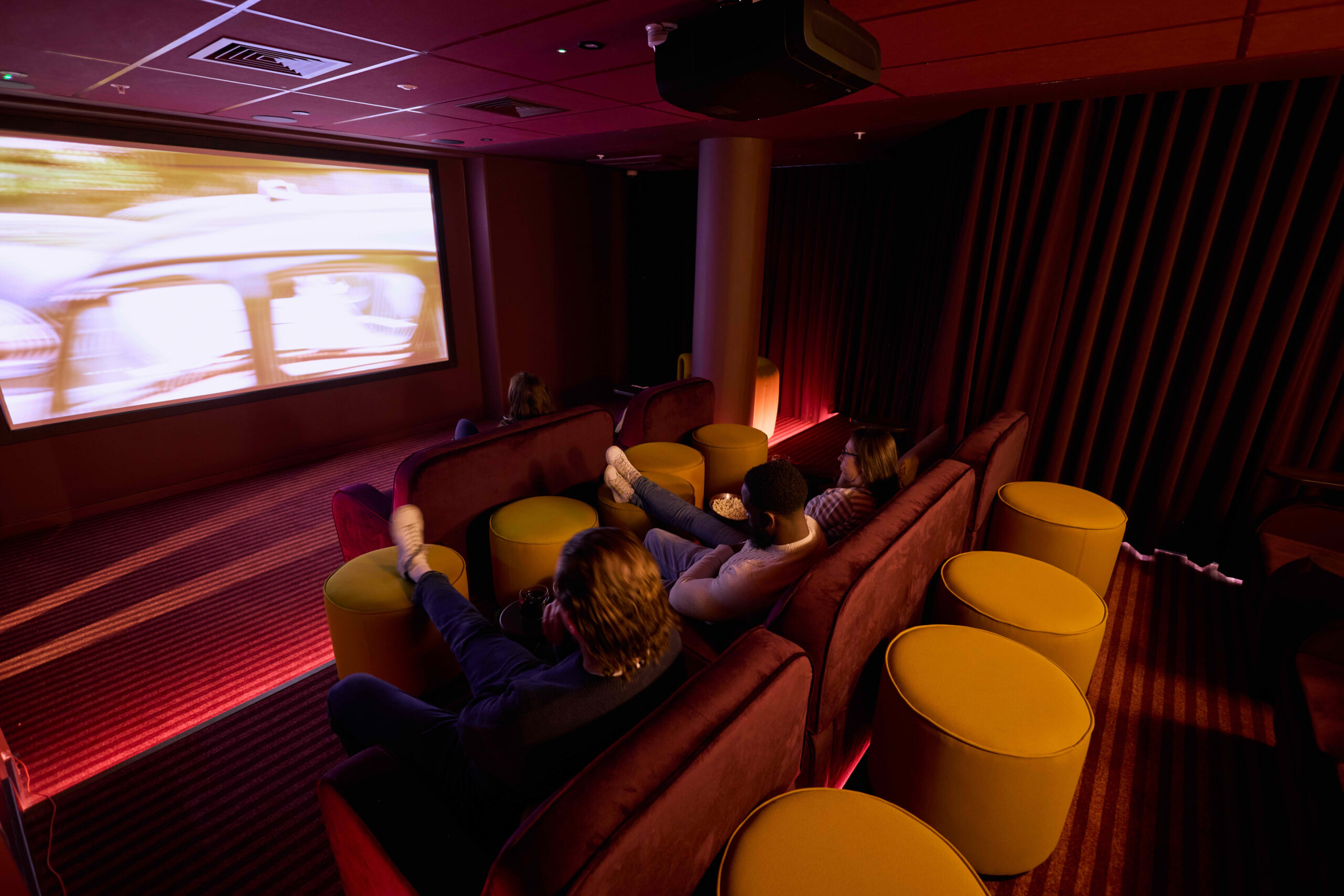 Luxury private cinema room at The Rex co-living in Kingston, London, with plush burgundy velvet seating and yellow footstools.