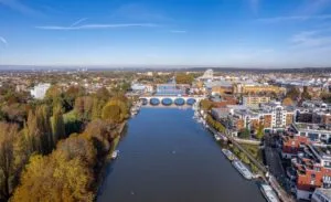 Overhead photograph of the River Thames in Kingston upon Thames, South West London, showing the river flowing under a multi-arched bridge. The location is captured in autumn, with rich yellow and brown foliage on the left bank and modern urban development, including riverside residential buildings and commercial areas, on the right bank under a clear blue sky.