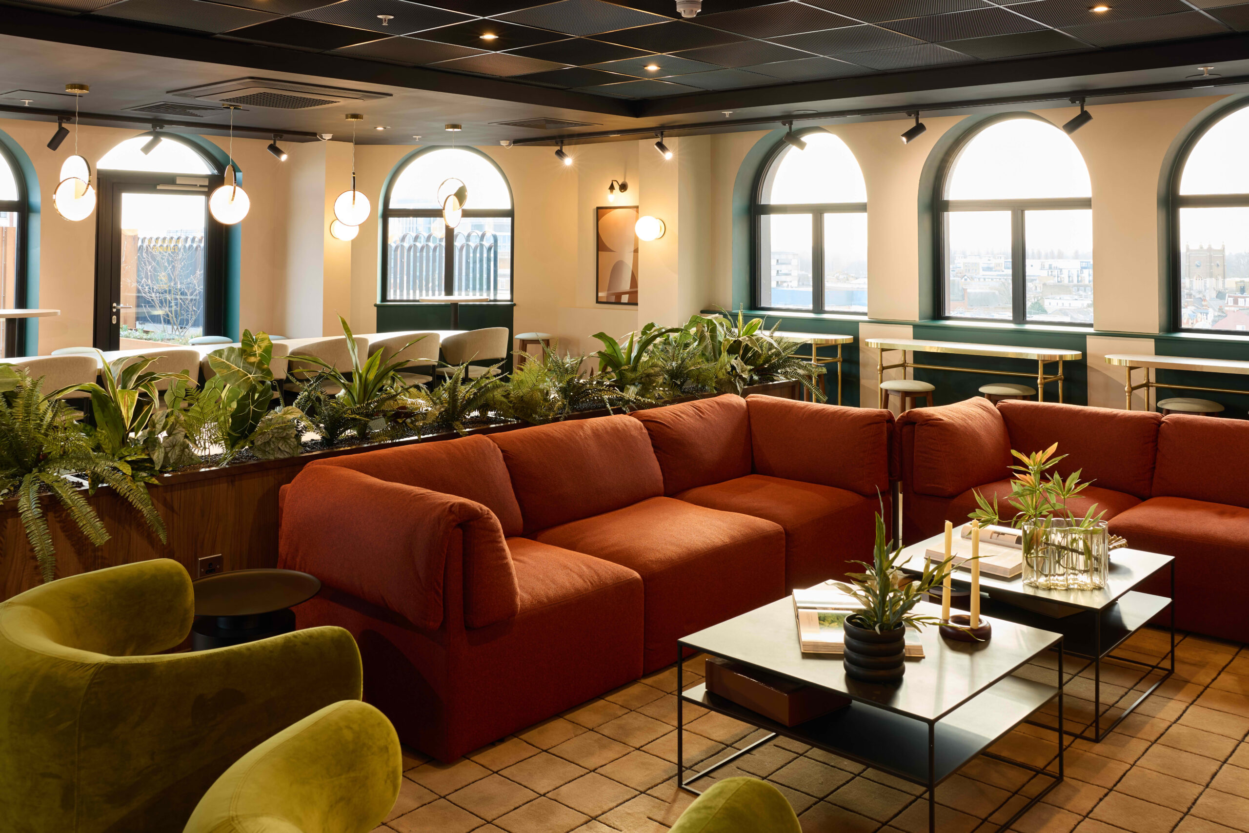 The expansive 7th-floor resident lounge at The Rex on Clarence Street, featuring mid-century modern green sofas, indoor plants, and floor-to-ceiling arched windows overlooking Kingston, London.