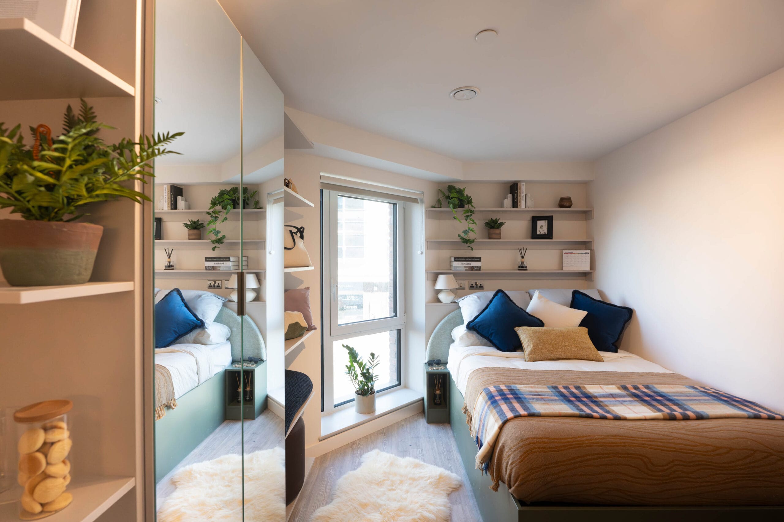 Bright, open-plan interior of a Comfort Type B studio at The Rex coliving, Kingston Upon Thames, featuring a set dining table, floor-to-ceiling mirrored storage, and a cosy double bed by a large window.